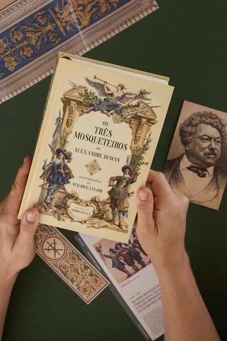 Mãos segurando a edição de Os Três Mosqueteiros ao lado do retrato colecionável de Alexandre Dumas